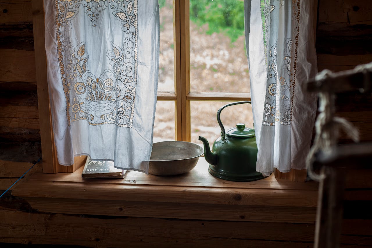 A cozy rustic windowsill with a green teapot and lace curtains overlooking the countryside.
